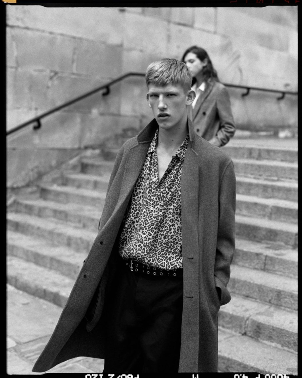 Immagine in bianco e nero di un uomo e una donna che scendono le scale. L'uomo indossa un cappotto a scacchi su una camicia con stampa leopardata e pantaloni neri.