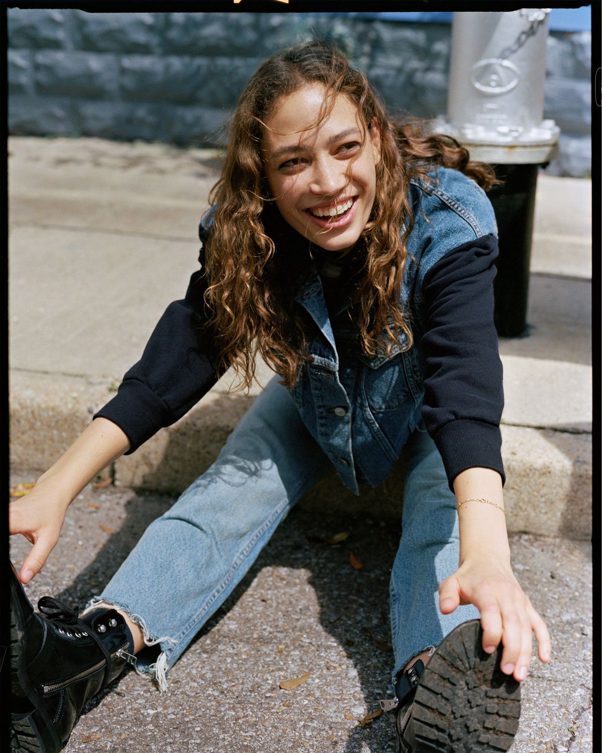 Divertida fotografía de una chica sentada en el asfalto alcanzándose la punta de los pies. Viste vaqueros de color azul índigo, botas negras de suela gruesa y una cazadora de denim de nuestra última colección.
