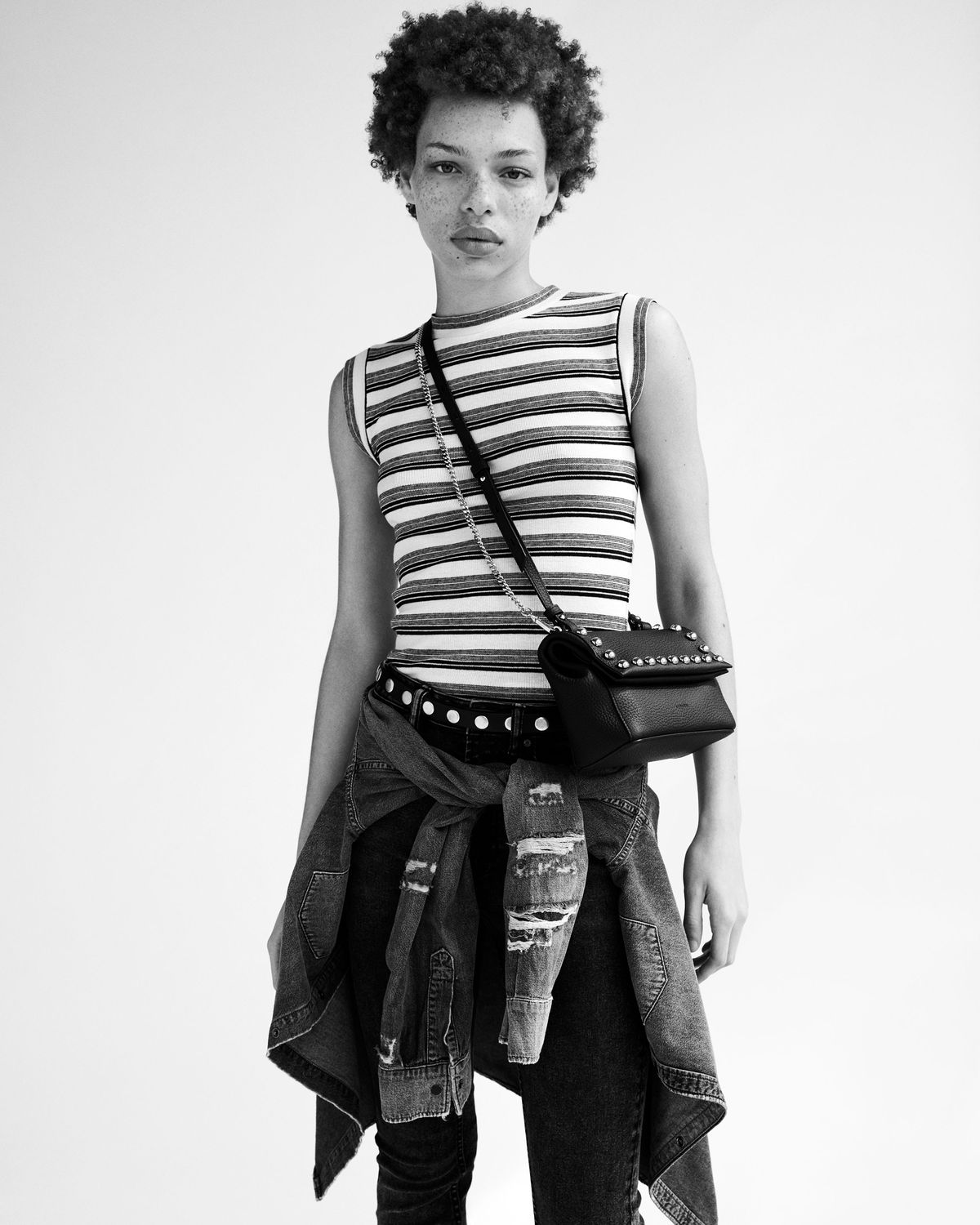 Black and white portrait of a young woman wearing a striped tank top and black jeans from our latest collection, complete with a studded leather crossbody bag and a denim jacket tied around the waist.
