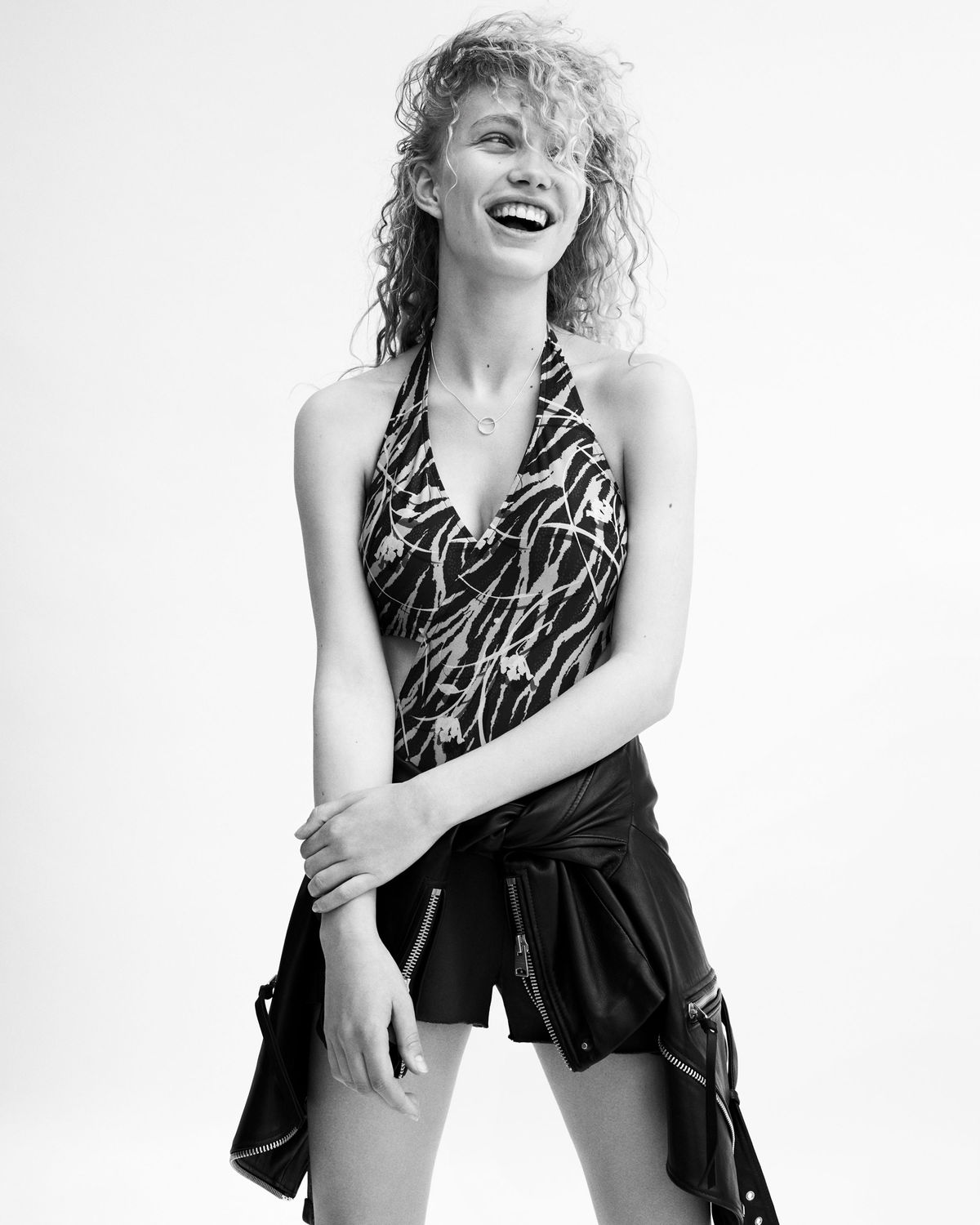 Black and white portrait of a young woman wearing a graphic swimsuit and black shorts, with a leather biker tied around the waist.