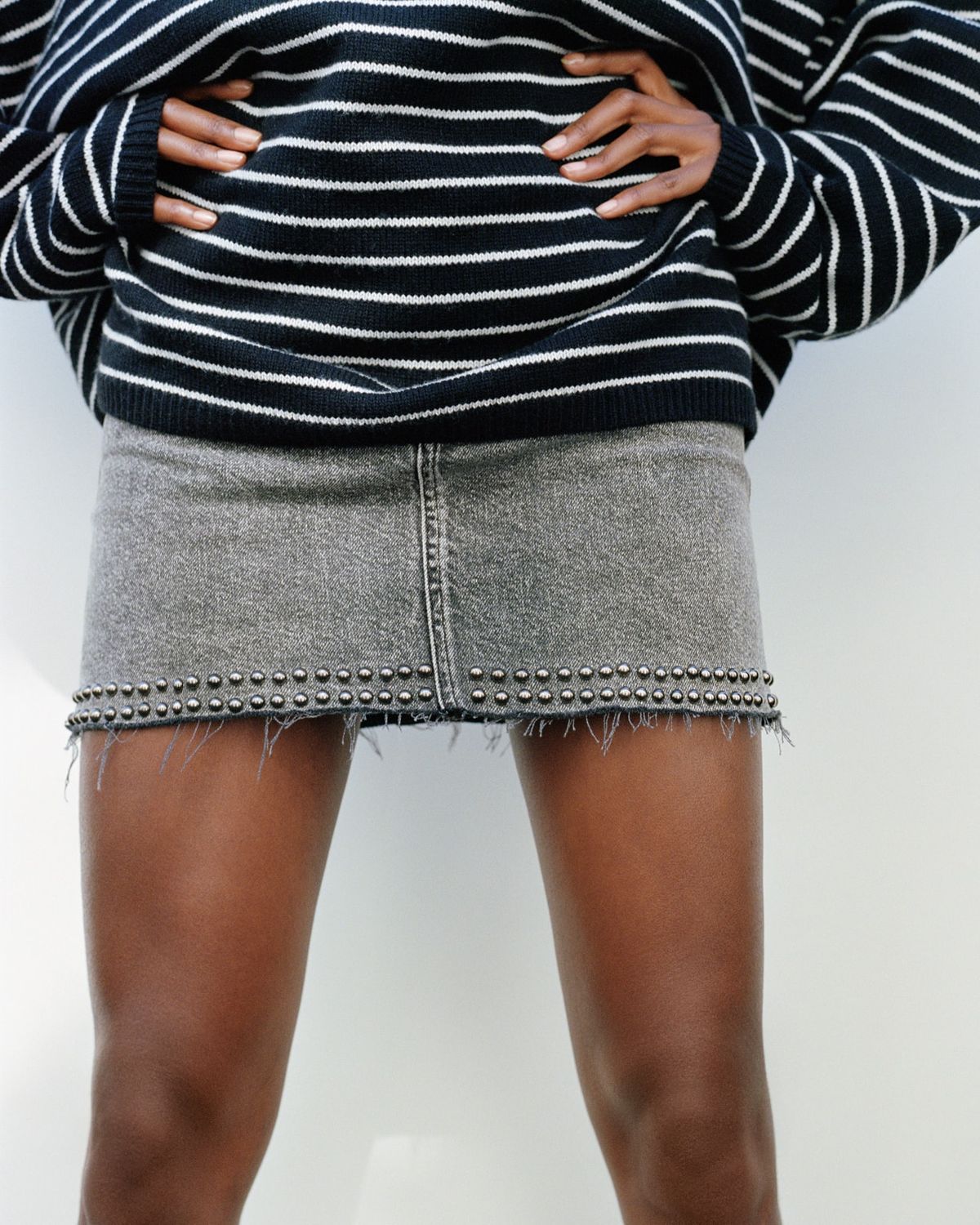 Image de campagne d'une femme portant une jupe en jean grise et courte à l'ourlet cloué avec un pull oversize rayé.