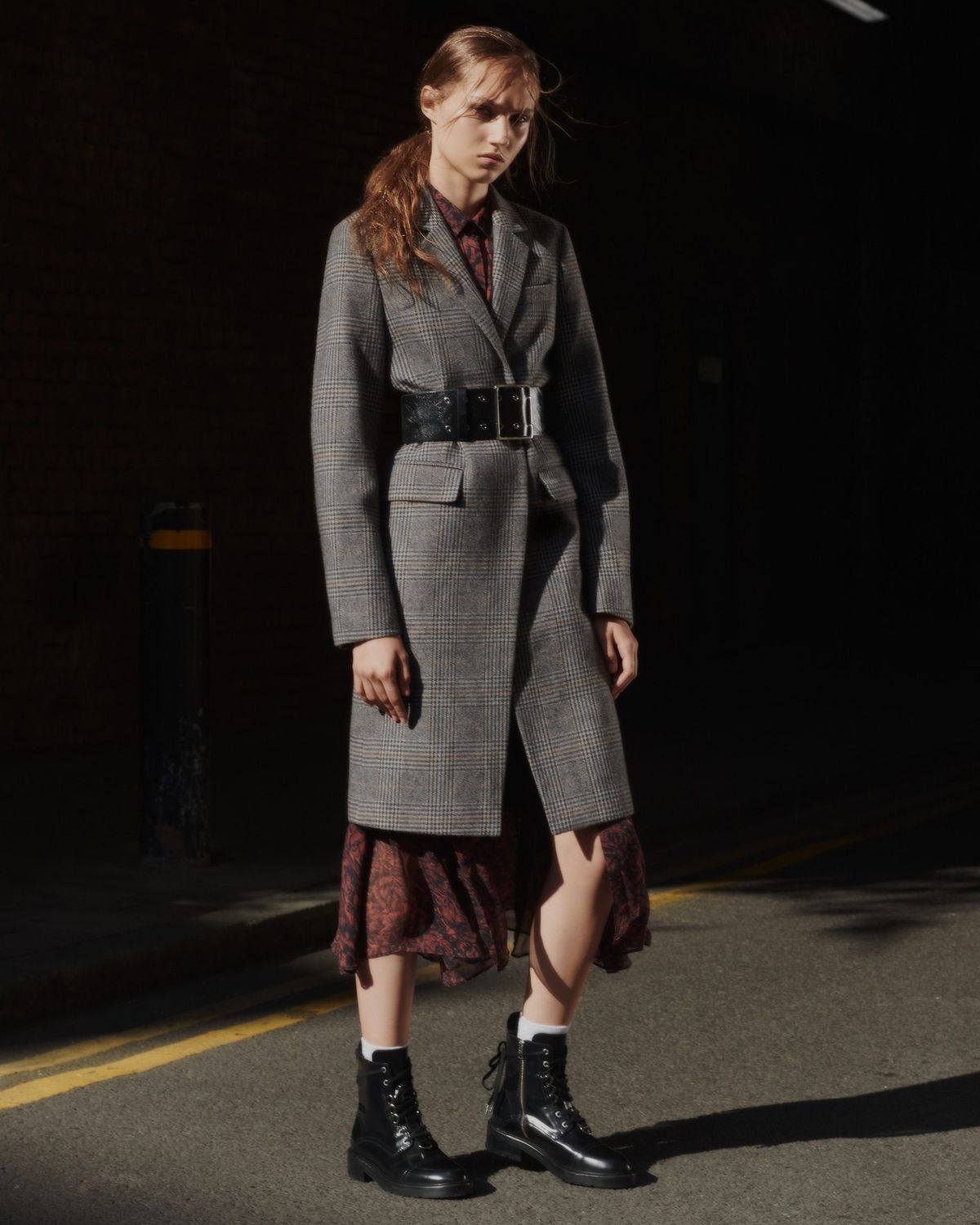 Image of a woman standing in the street wearing a check coat, styled with a large belt at the waist and military boots, over a long flower printed red dress.
