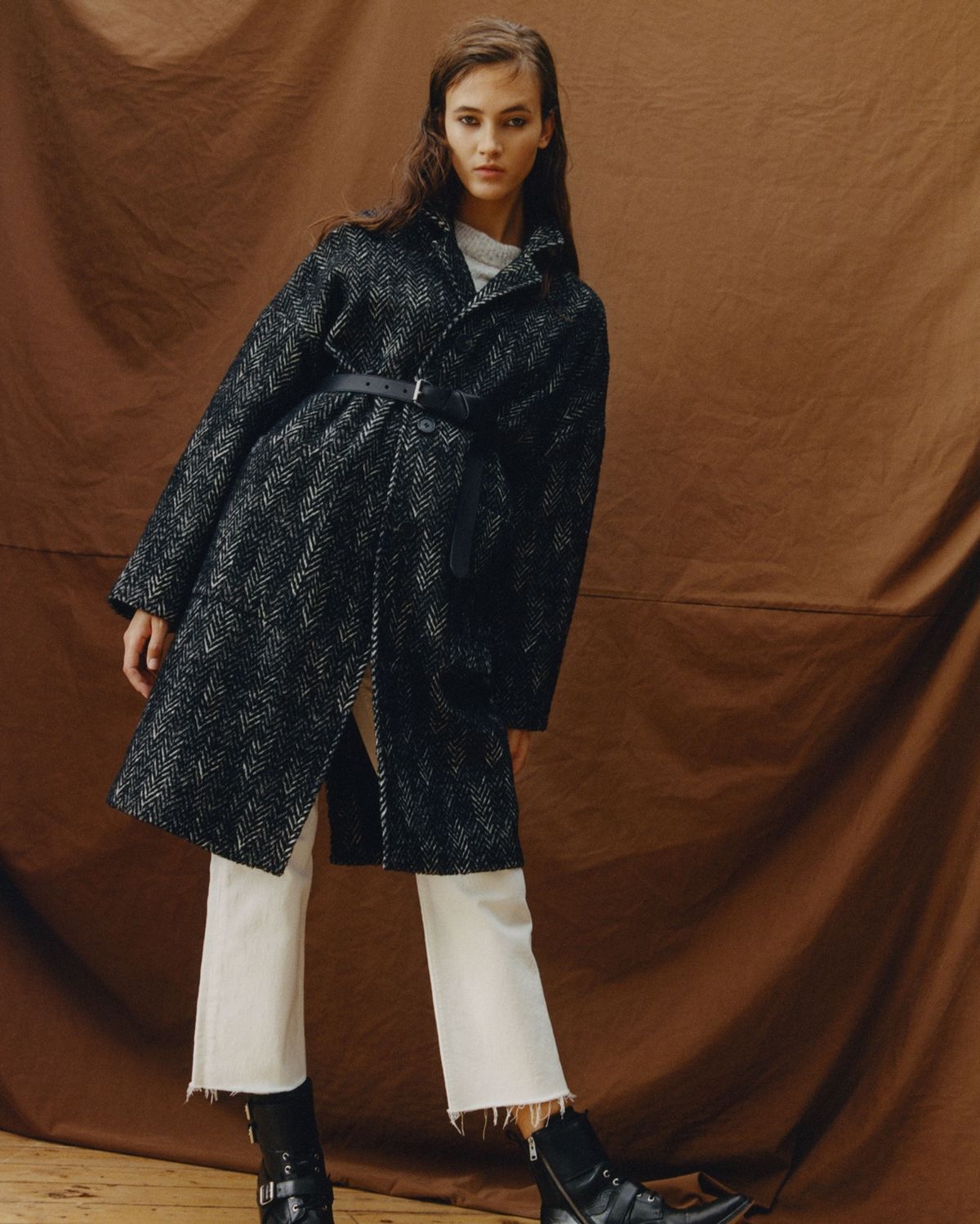 Image of a woman wearing a black and white Herringbone coat, closed up with a black leather belt, with white jeans and black boots.