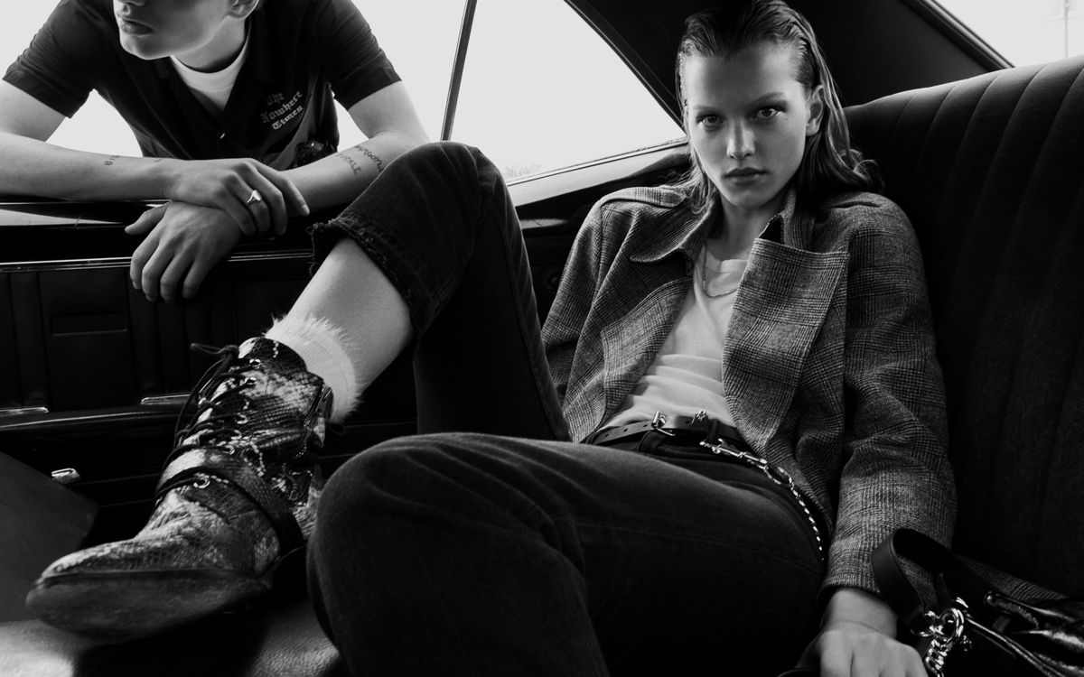 Image of a woman sitting at the back of a car wearing a checked mac with white t-shirt, black jeans and snake printed boots. A man leaning at the car window wears a black short sleeve shirt on top of a white t-shirt.