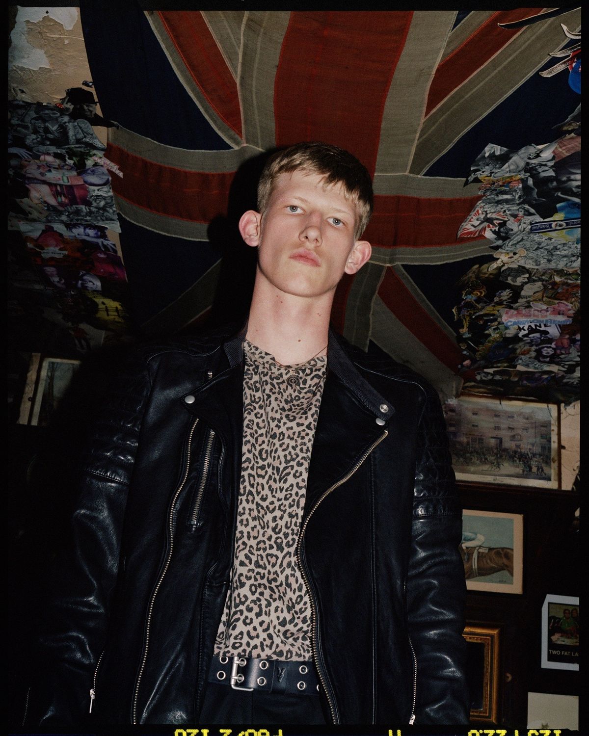 Kampagnenfoto von einem Mann, der vor einem Hintergrund voller Erinnerungsstücken aus dem Vereinigtem Königreich steht und eine schwarze Lederjacke über eine Leoprint T-Shirt trägt.