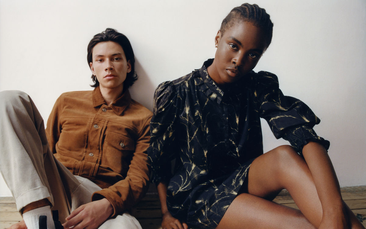 Man and a woman siting on the floor in front of a white wall wearing items from our new collection.