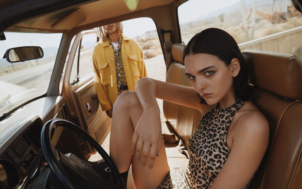 Image of a woman seating behind the wheel of a car and a man standing by the open door, both wearing items of our new collection.