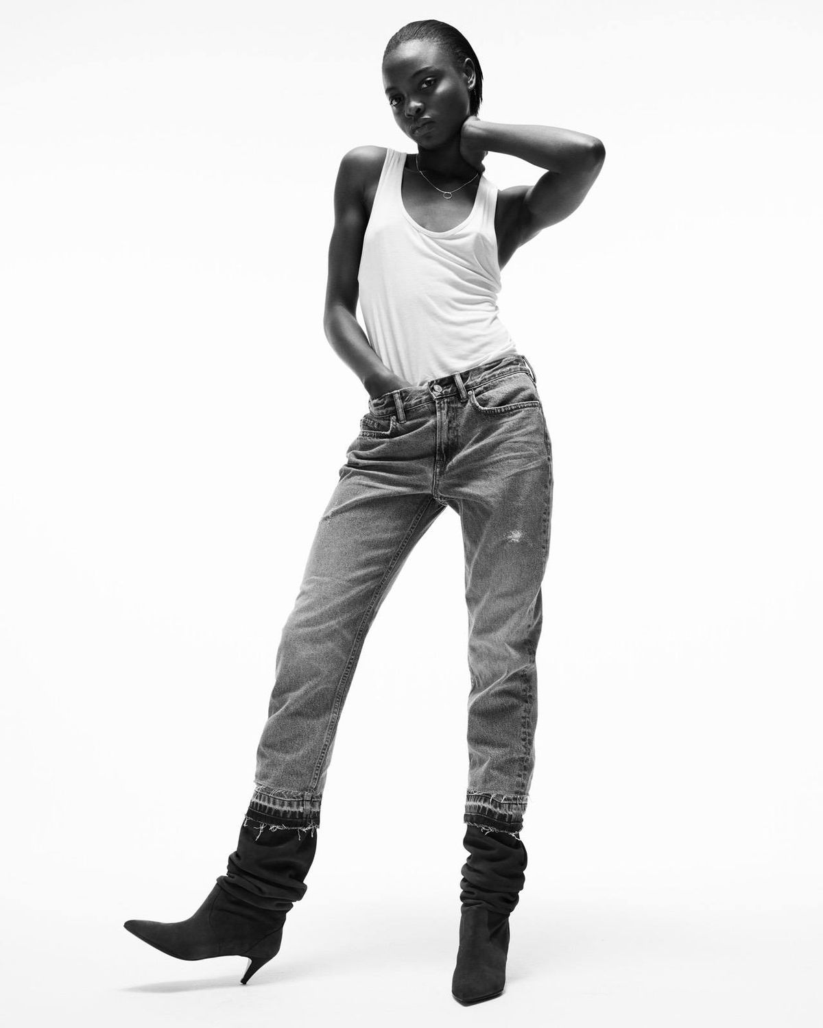 Black and white image of a woman wearing a white tank top with light straight jeans and 80s inspired slouch boots.