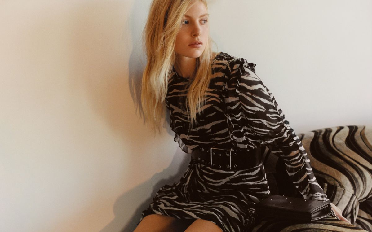 Image of a woman sitting on a couch and wearing a long sleeve zebra printed dress with a wide leather belt at the waist and a black studded clutch bag.