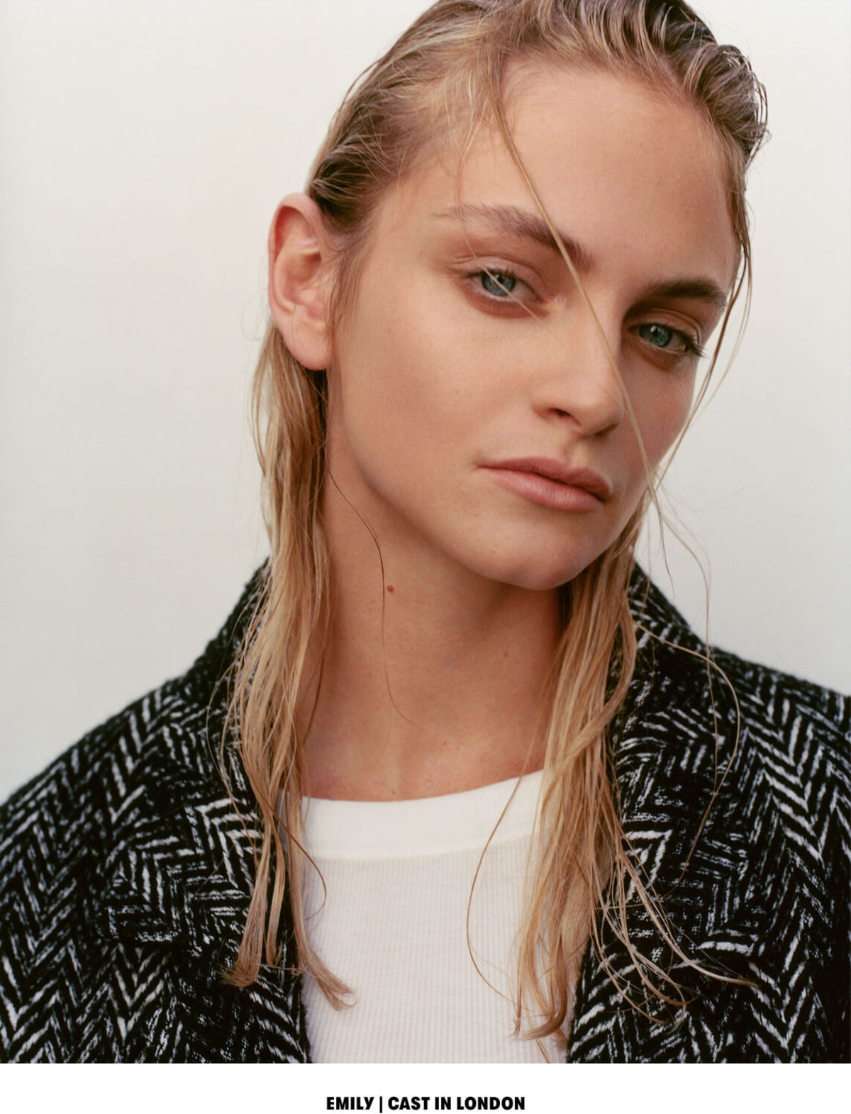 EMILY | CAST IN LONDON. Close up portrait of a blonde woman wearing a herringbone coat over a white t-shirt.