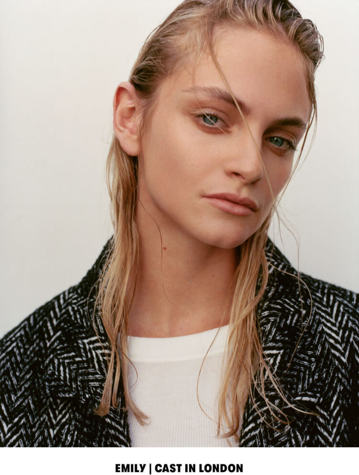 EMILY | CAST IN LONDON. Close up portrait of a blonde woman wearing a herringbone coat over a white t-shirt.