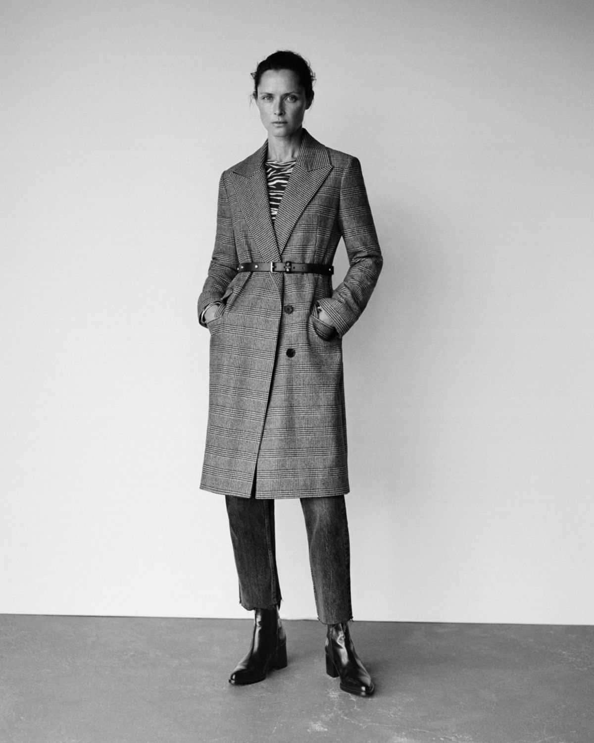 Black and white image of a woman standing in a photo studio and wearing a long checked tailored coat over a zebra printed t-shirt and dark denim jeans.
