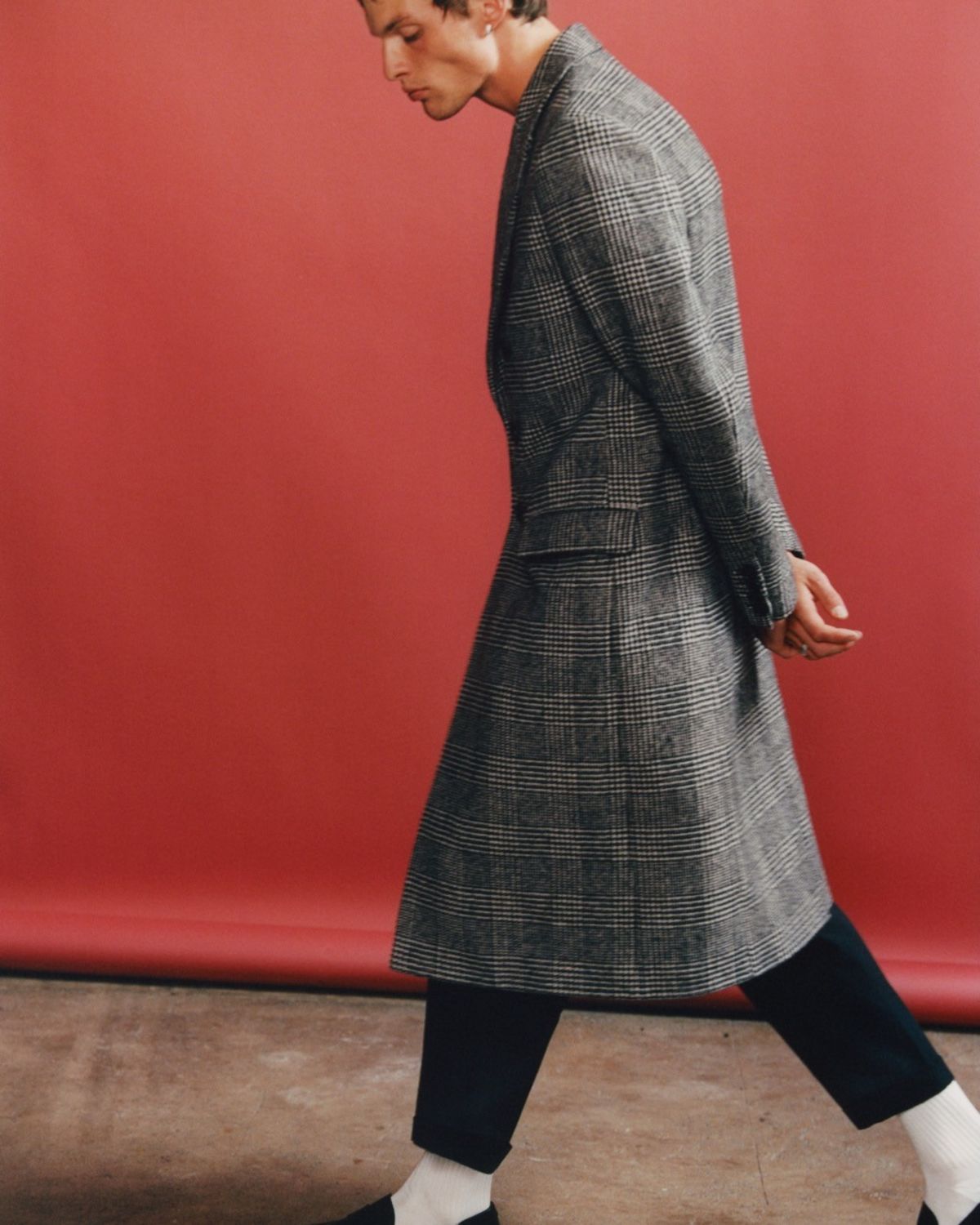 Image of a man walking in front of a red background wearing a long grey checked coat with black tailored trousers.