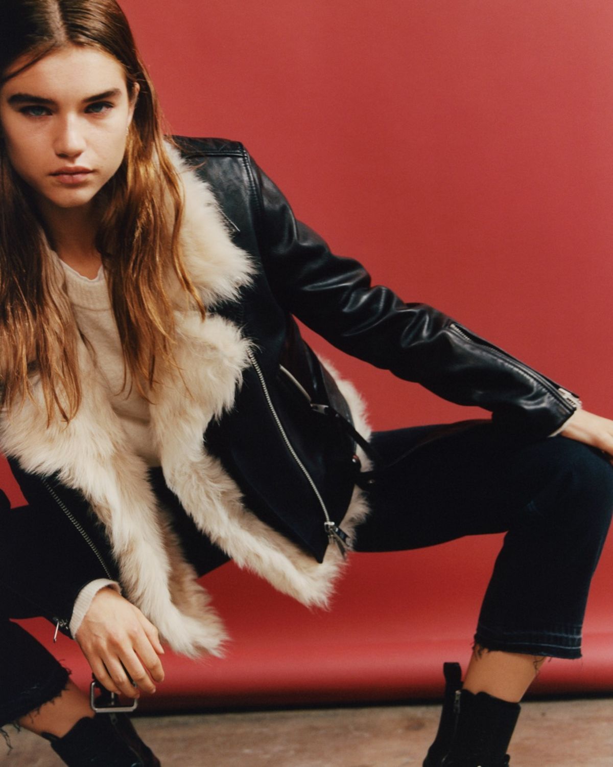 Image of a woman crouching in front of a red background and wearing a black leather jacket fur a white fur lining with a white jumper, black jeans and black boots.