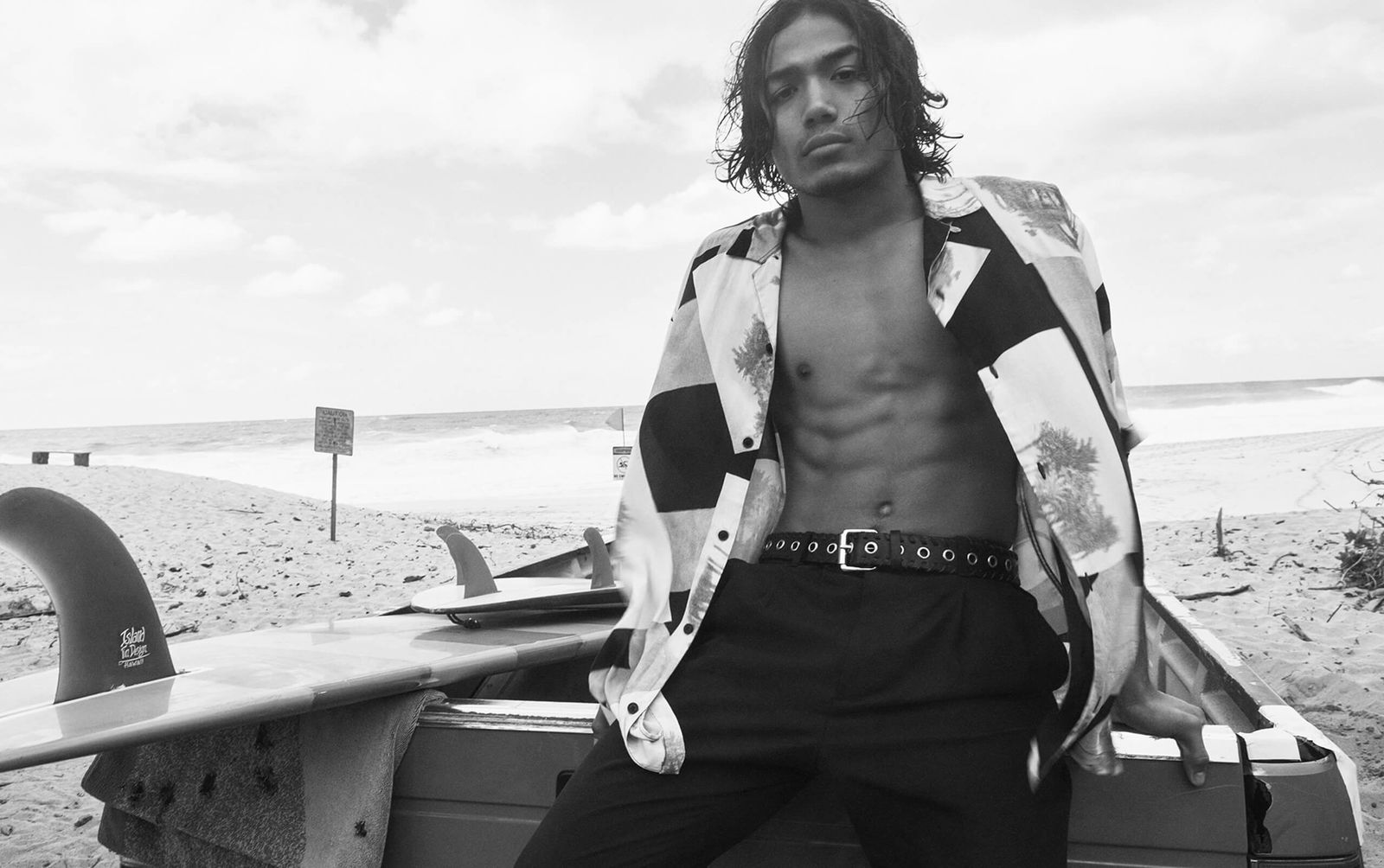 Black and white landscape image of a man sitting at the back of a pick up truck, wearing an opened printed Hawaiian shirt with black tailored trousers.