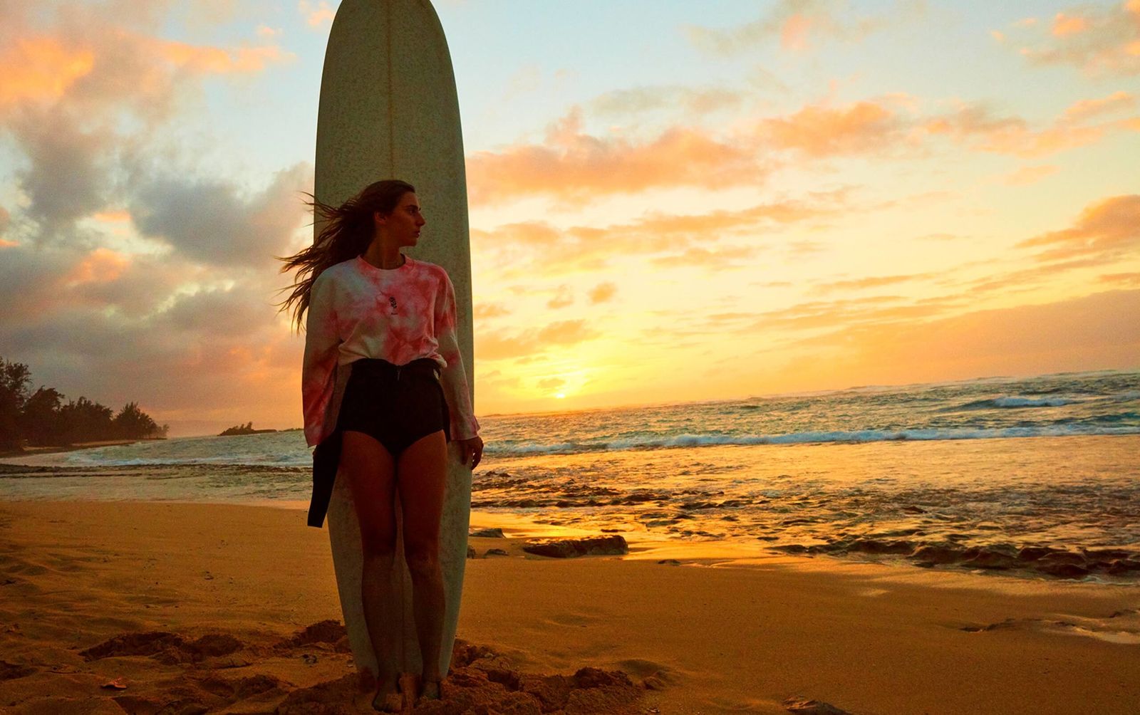 Photo paysage d'une femme debout appuyée à sa planche de surf au coucher du soleil.