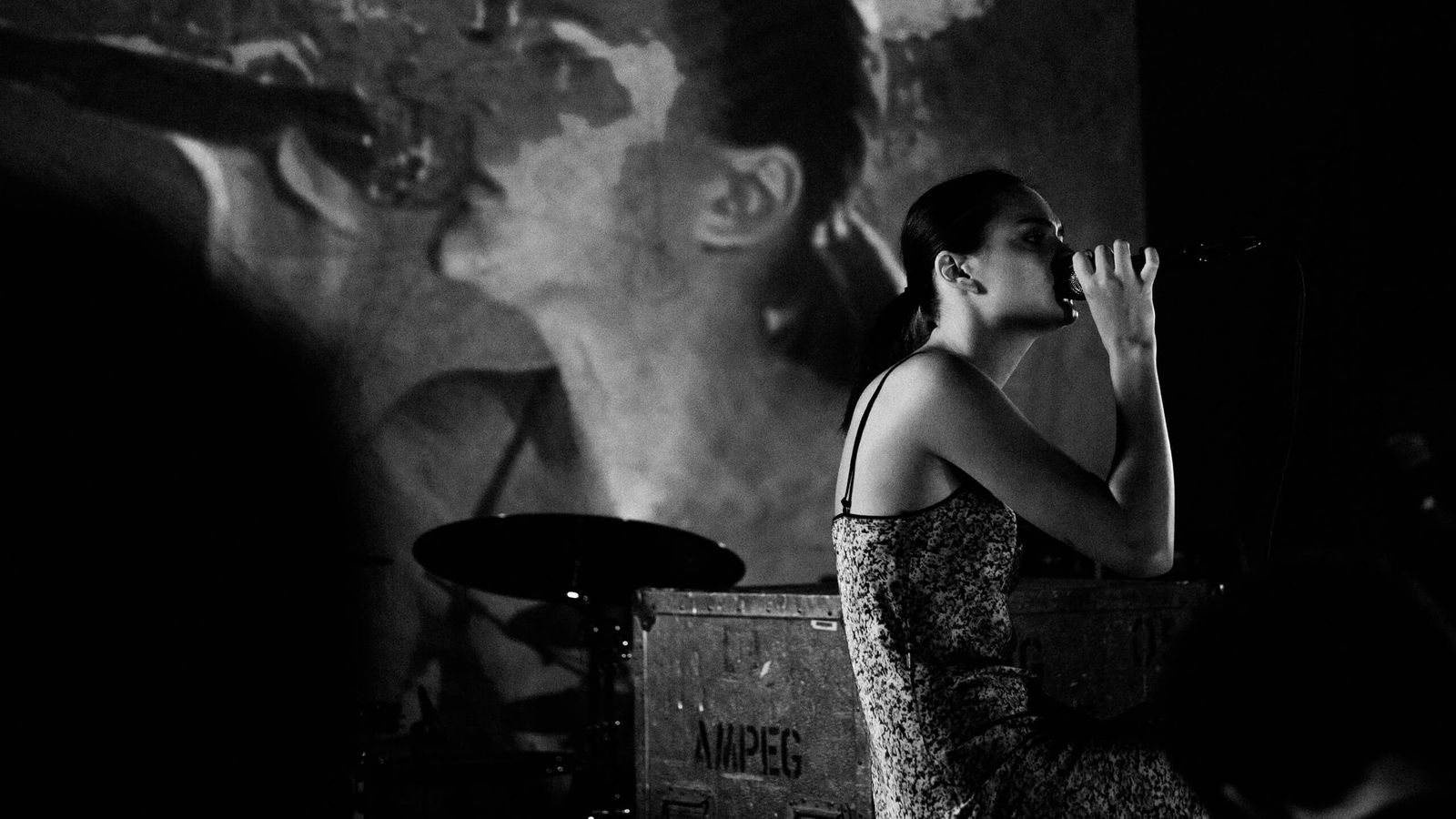 Black and white image of Charlotte Lawrence sitting on stage and singing, wearing a thin strap flower printed dress.