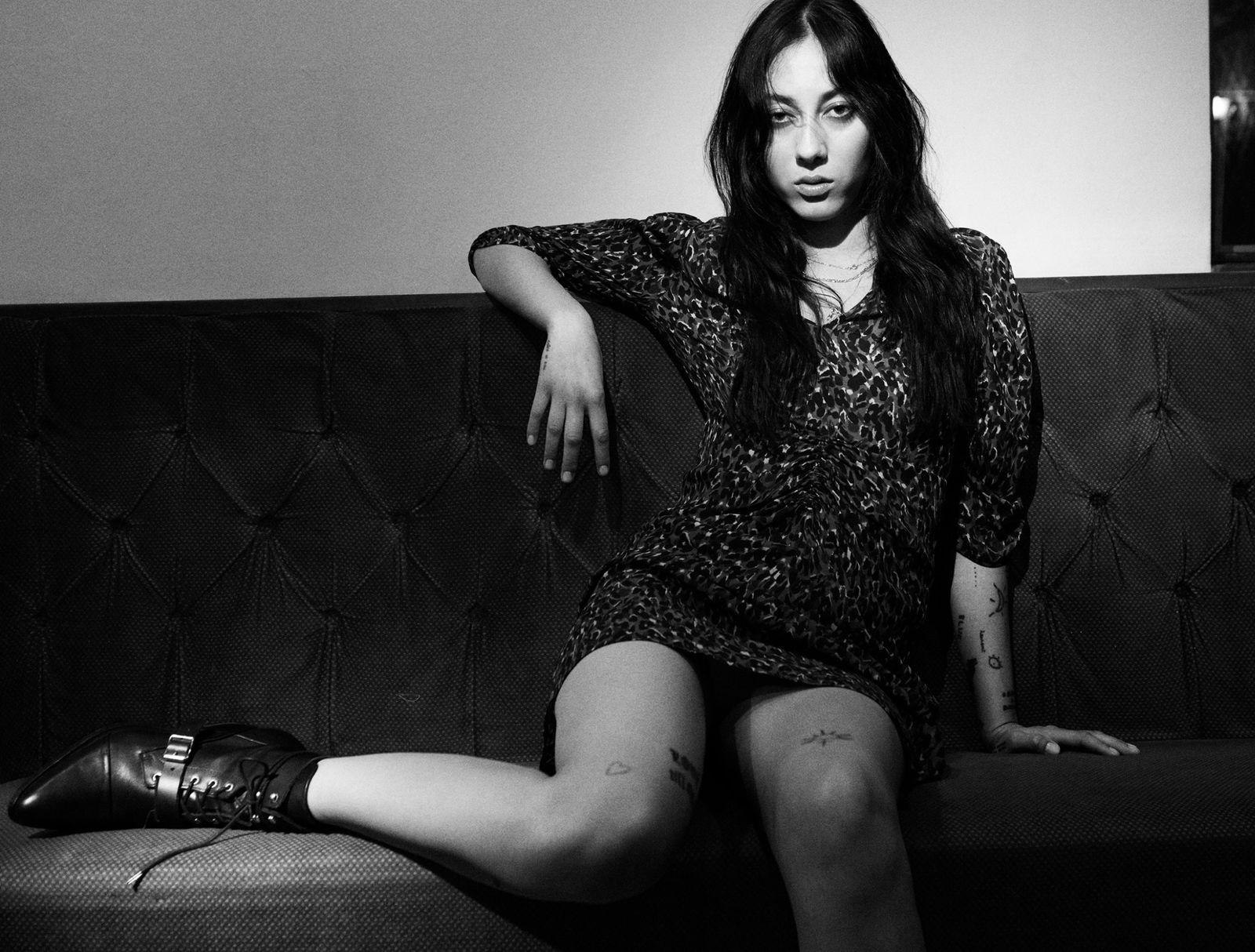 Black and white image of a woman sitting on a couch wearing a short leopard printed dress with black leather boots.