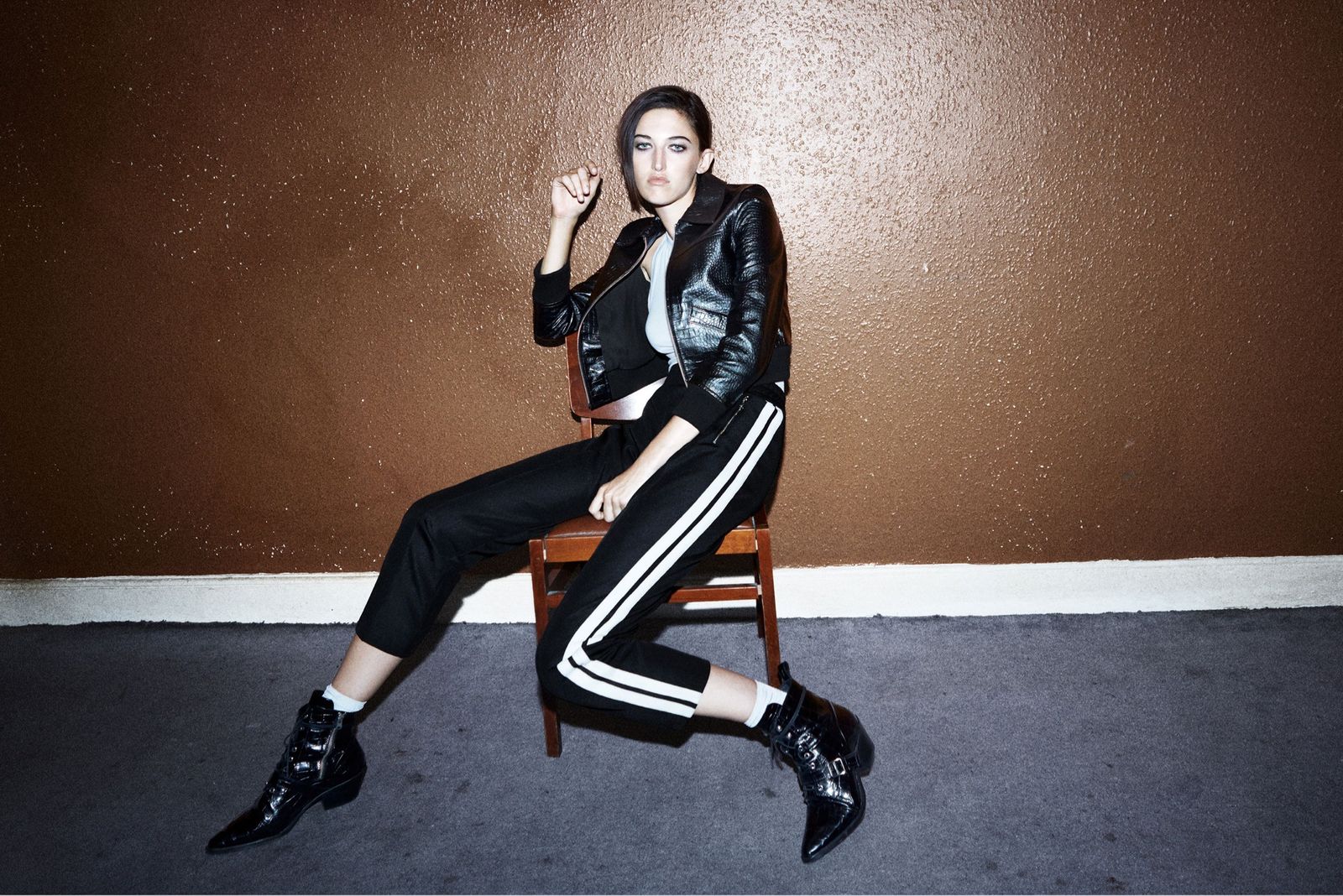 Image of a woman sitting on a chair wearing a cropped leather jacket with black trousers with white stripes down the side and black croco boots.