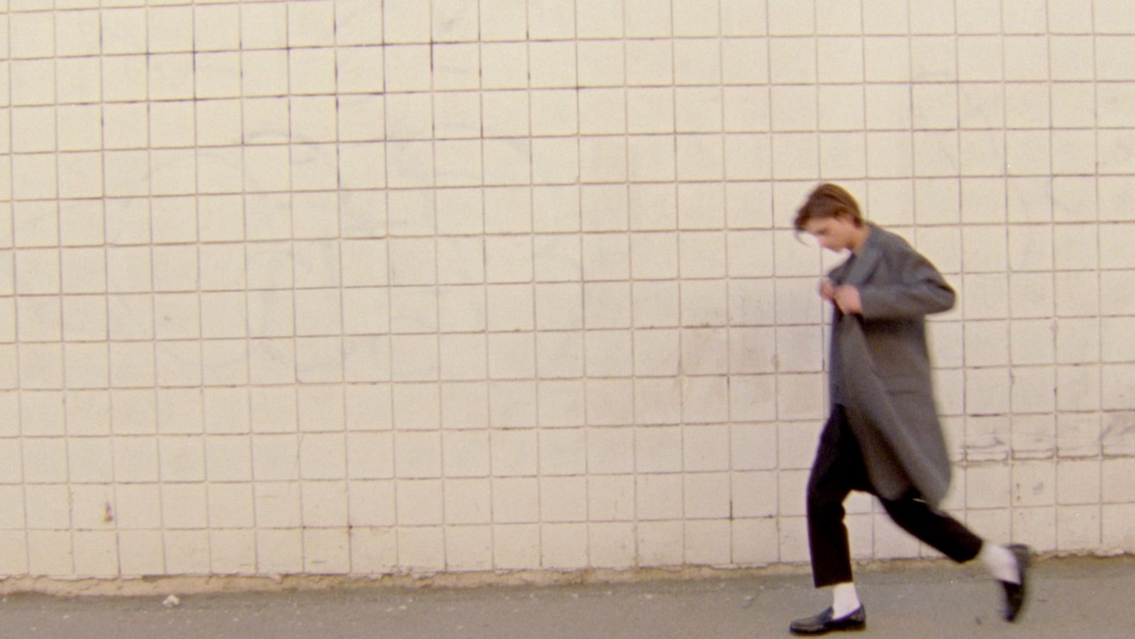Mini-clip screenshot image of a man walking in a street wearing black tailored trousers with black loafers and a long grey coat.