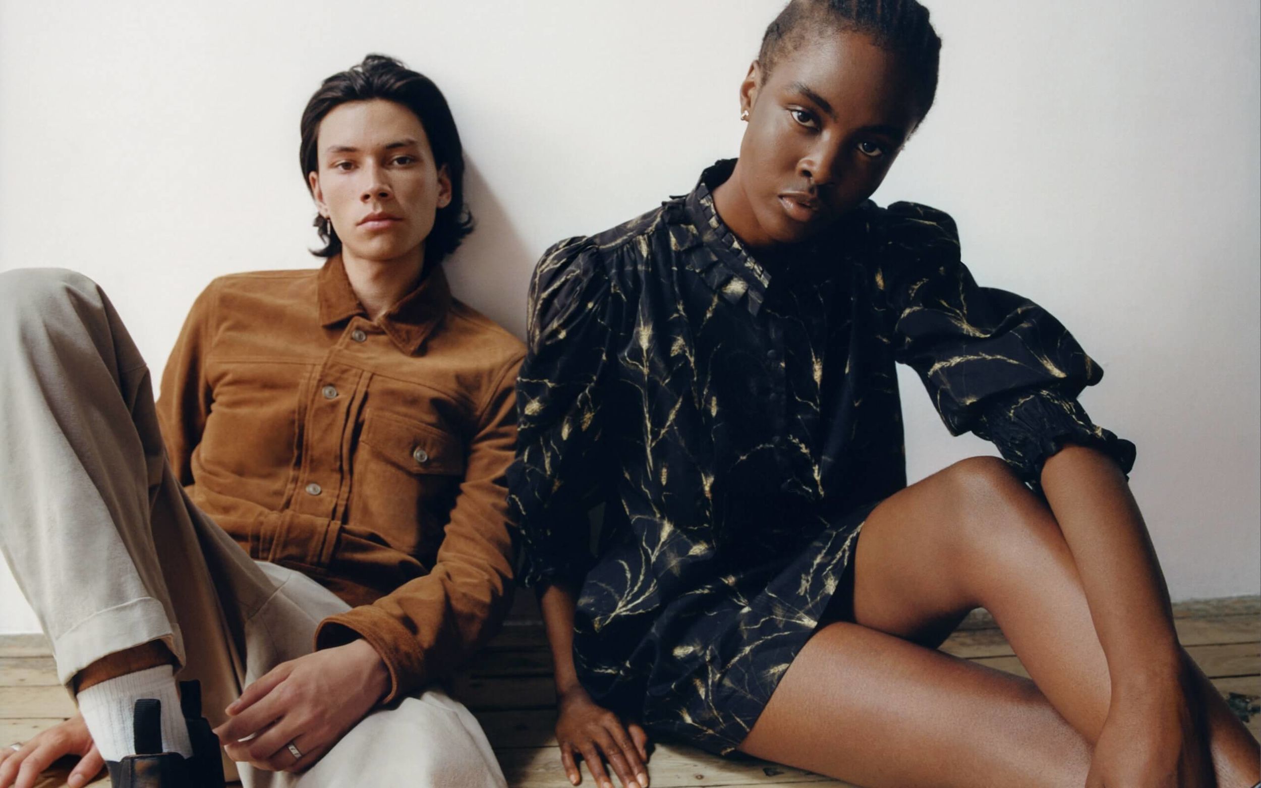 Woman and a man sitting on the floor against a wall wearing items from our latest collection.