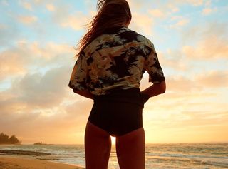 Photo d'une femme de dos regardant la mer et portant une chemise hawaïenne sur un maillot noir.