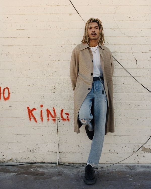 Man standing against a wall wearing light denim jeans with a white t-shirt and beige raincoat.