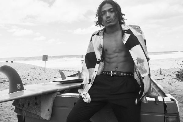 Black and white picture of a man sitting at the back of a truck wearing an opened hawaiian shirt with a leather belt and black trousers.