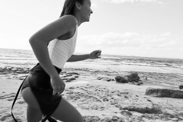 Black and white image of a woman running on the beach wearing a white swimsuit and a wetsuit.