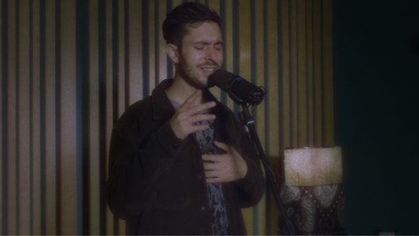 Khushi debout à son micro, interprétant sa chanson. Il porte un pull gris à motif léopard et une surchemise foncée.