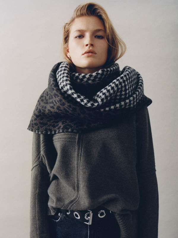 Woman standing wearing a dark grey jumper with an obersize printed scarf around her neck.