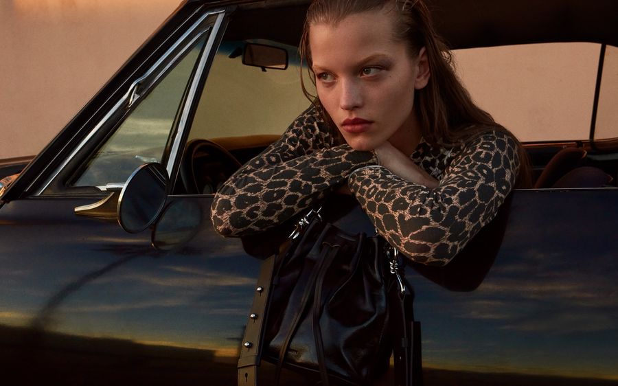 Ritratto di una donna seduta sul desile di una macchina sportiva nera. Indossa una maglia a maniche lunghe con stampa leopardata e porta una borsa nera in pelle.