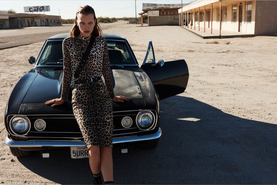 Immagine di una donna appoggiata ad una macchina nera sportiva. Indossa una gonna leopardata e una t-shirt in coordinato con una borsa in pelle nera e un paio di stivali neri.