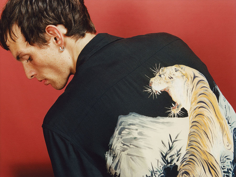 Foto di un uomo di schiena di fronte ad un muro rosso. Indossa una camicia hawaiana nera con una tigre sul retro.