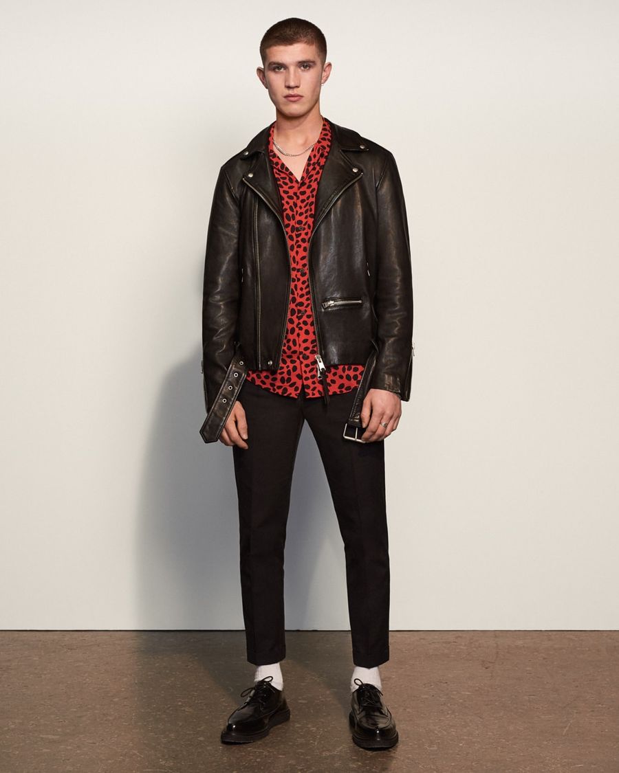 Foto von einem Mann, der eine schwarze Lederjacke, ein rot und schwarz gepunktetes Hemd und eine schwarze Hose trägt.