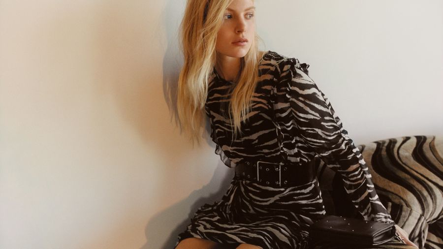 Image of a woman sitting on a couch and wearing a long sleeve zebra printed dress with a wide leather belt at the waist and a black studded clutch bag.