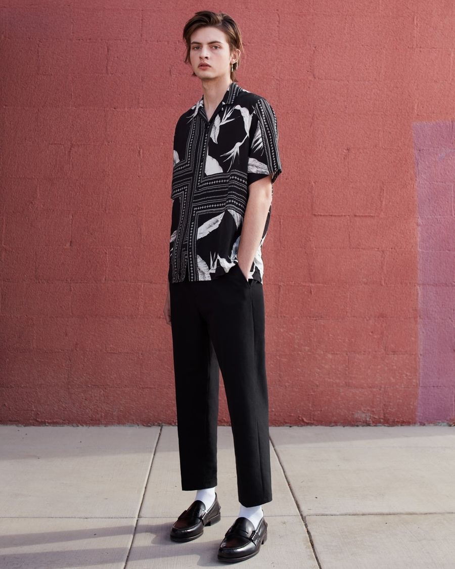 Image of a man standing in front of a pink wall and wearing a black and white printed shirt with black tailored pants.