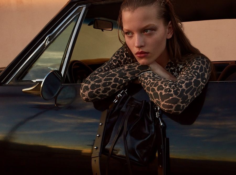 Portrait of a woman sitting in a black sport car wearing a long sleeve leopard print top and holding a black leather bag.