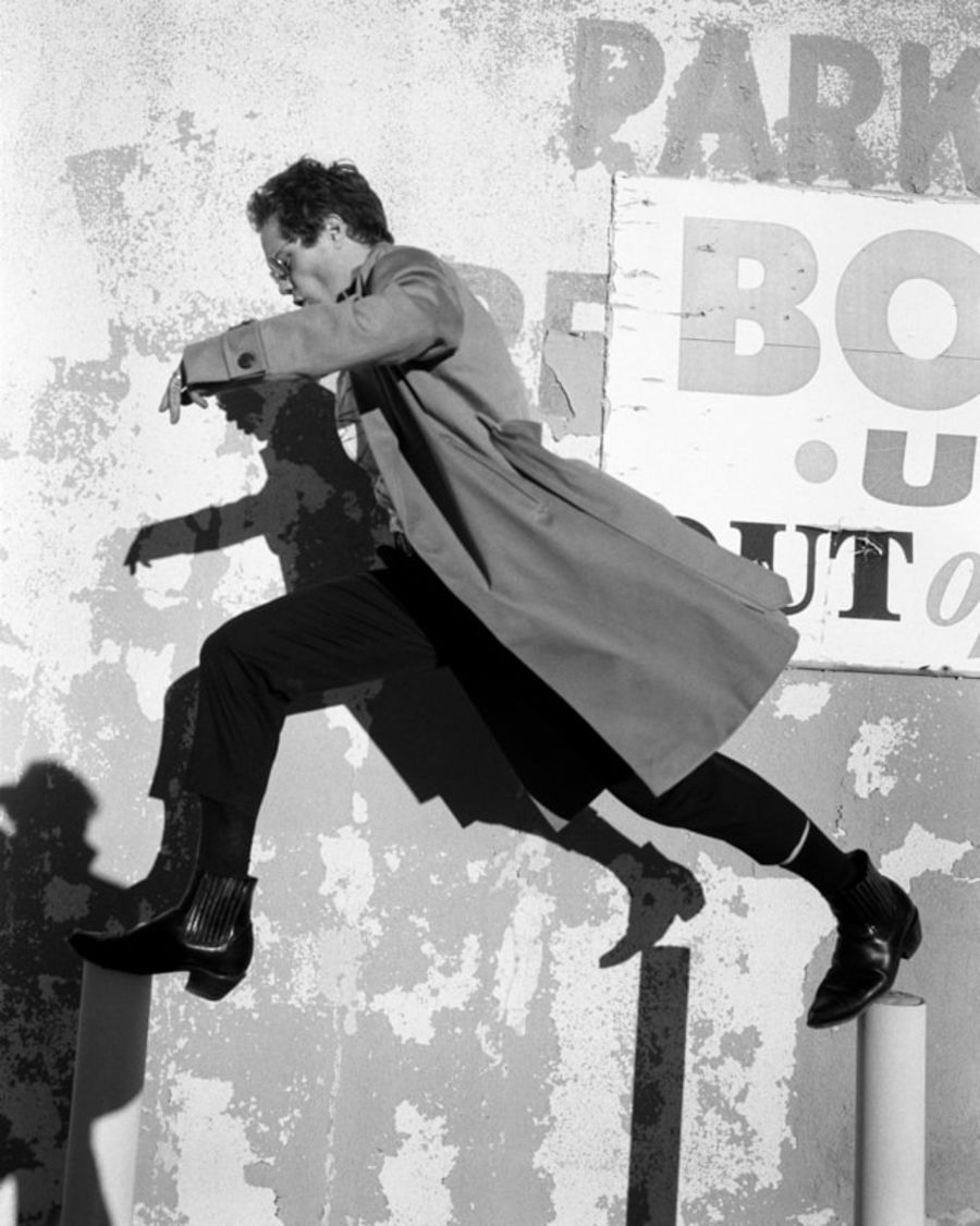 Black and white image of a man jumping in the street wearing black tailored trousers, black western boots and a camel trench coat.