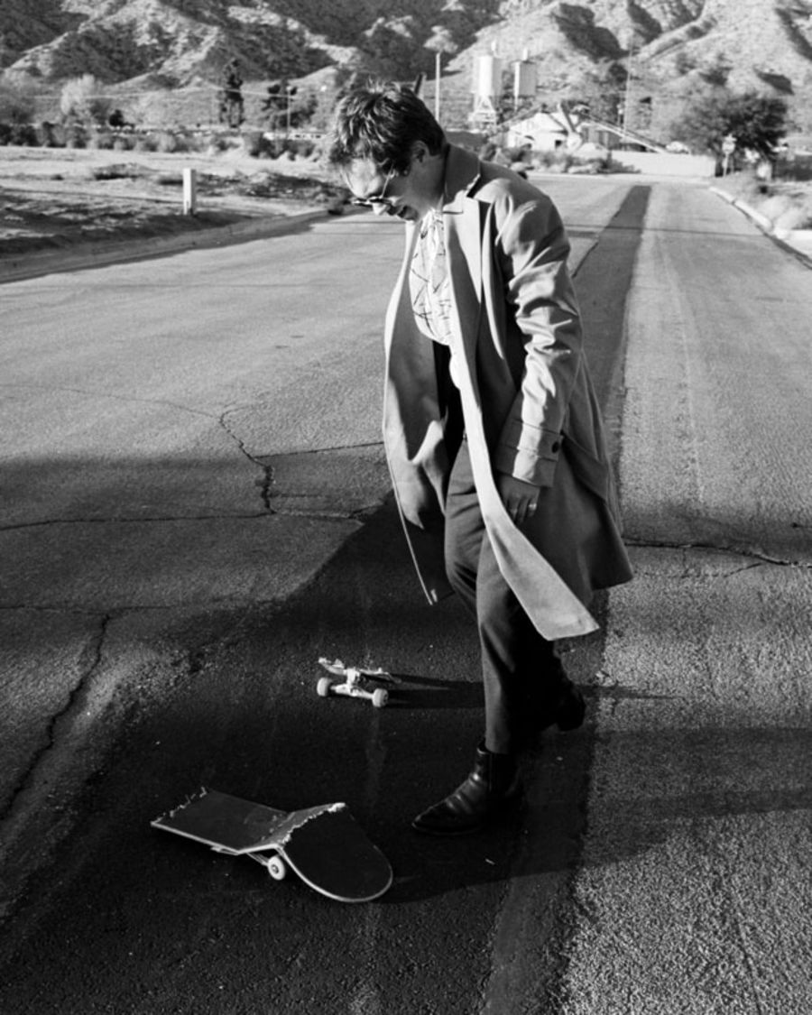Black and white image of a man wearing pieces from our latest collection and looking down at a broken skate board.