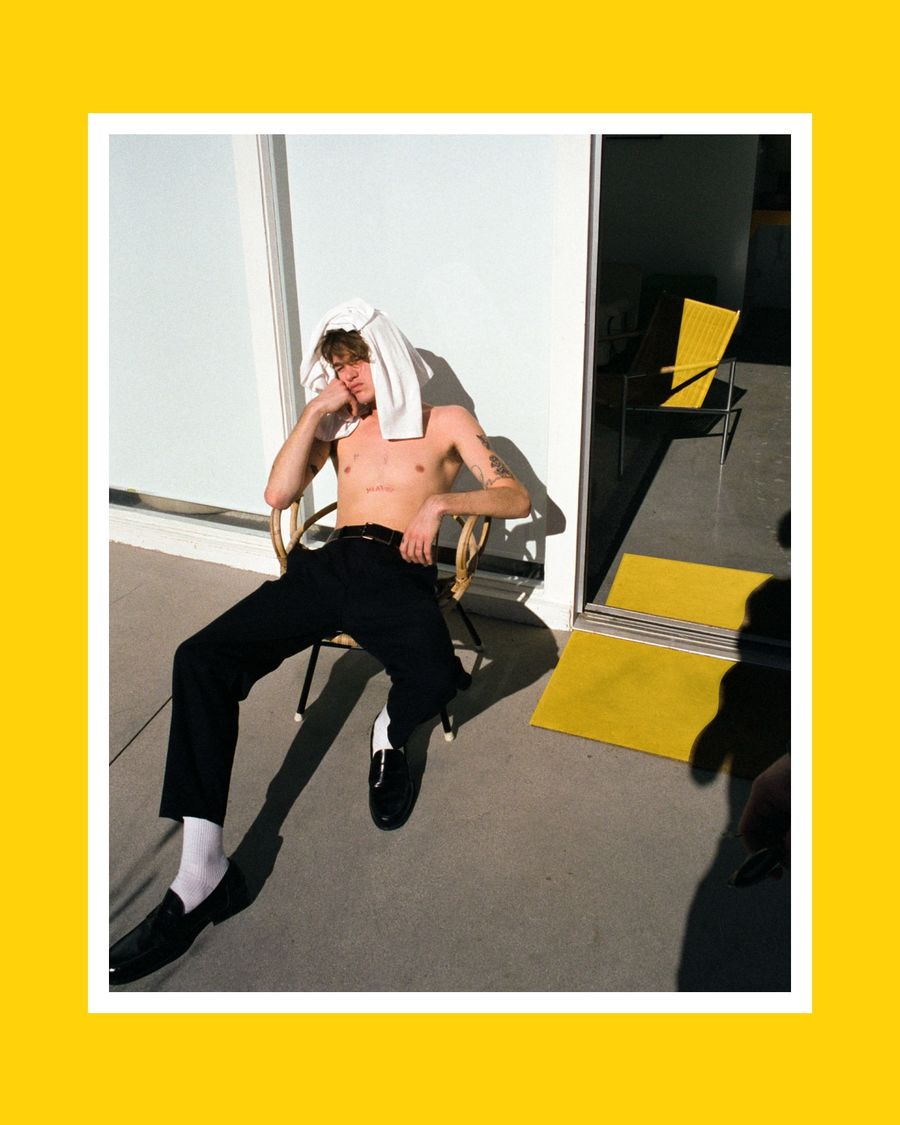 Image of a man sitting on a chair in the sun wearing black tailored trousers and a t-shirt on his head.