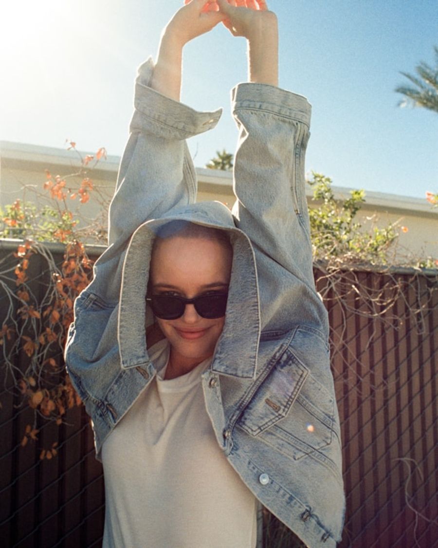 Portrait of a woman with her arms up in the air wearing a white tank top with a light denim jacket.