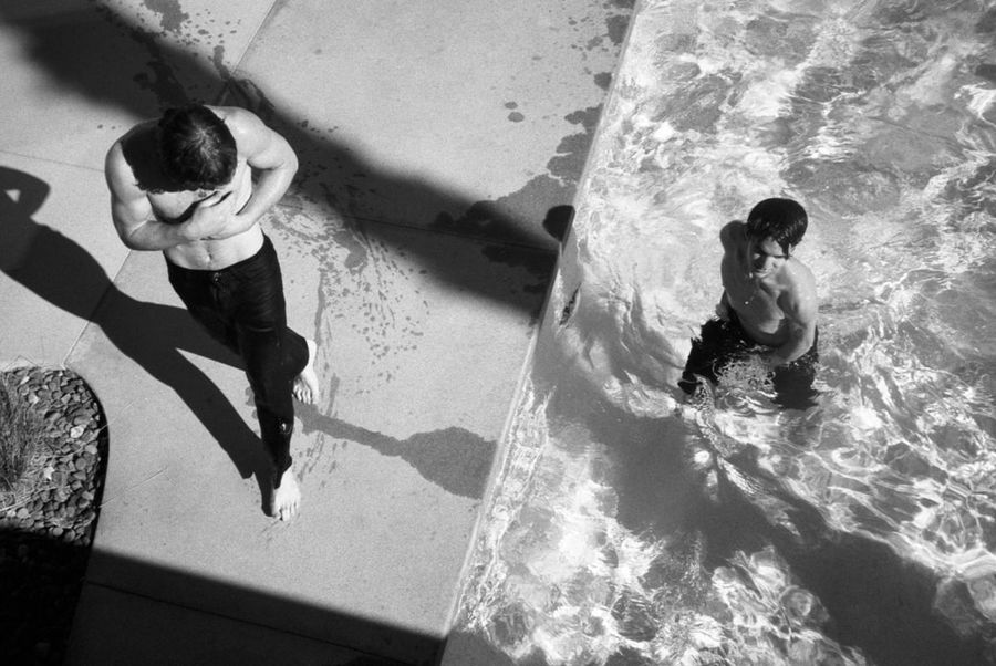 Black and white image of two men in a swimming pool wearing black tailored trousers.