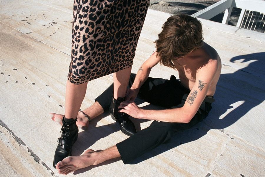 A man sitting on the floor wearing black tailored trousers is lacing the boots of a woman standing and wearing a midi leopard skirt with black leather shoes.