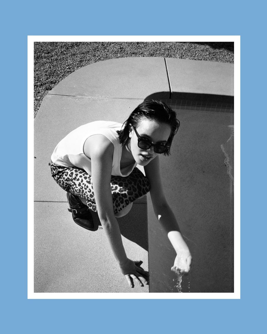 Black and white image of a woman crouched down near a pool and wearing a leopard skirt with white tank top.