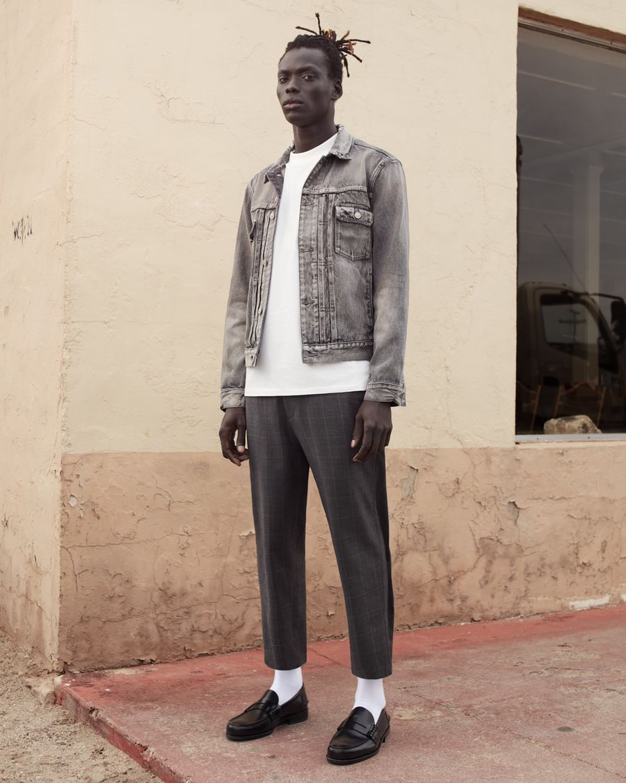 Foto von einem Mann, der vor einer hellgelben Wand steht und eine hellgraue Jeansjacke mit einem weißen T-Shirt, grau karierten Hosen und schwarzen Loafern trägt.
