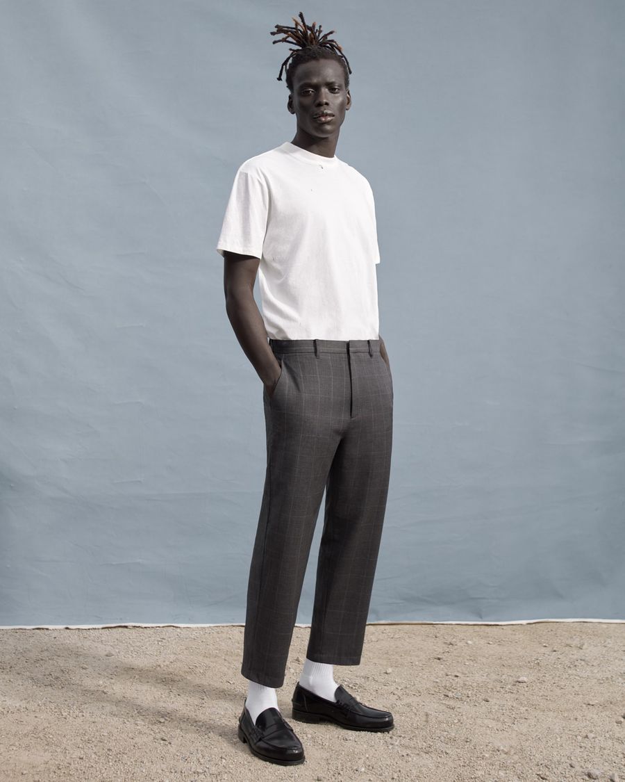 Foto von einem Mann, der vor einer grauen Wand steht und ein weißes T-Shirt, eine graue karierte Hose und schwarze Loafer trägt.