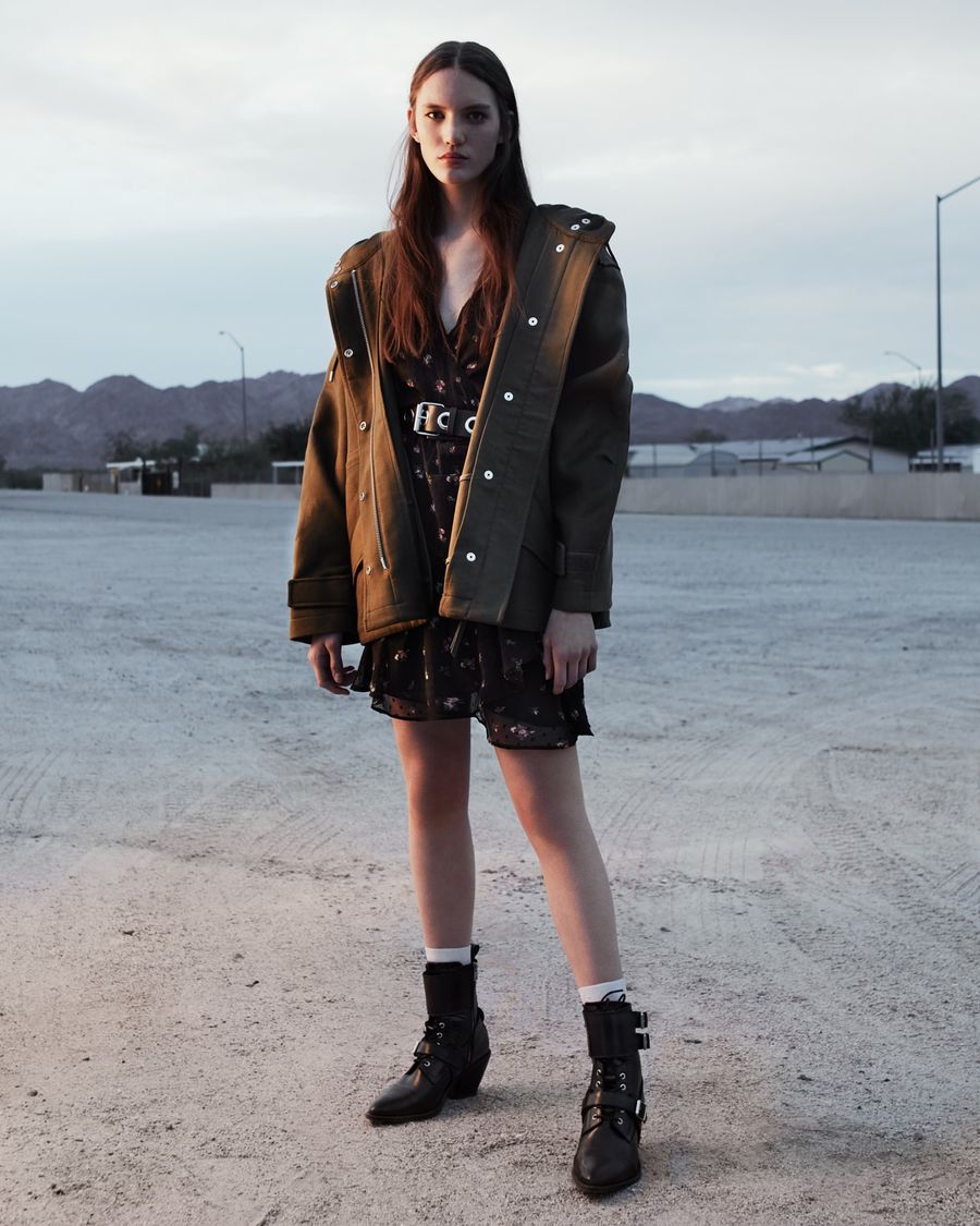 Image of a woman standing outside wearing a khaki jacket opened over a short printed dress and leather boots.