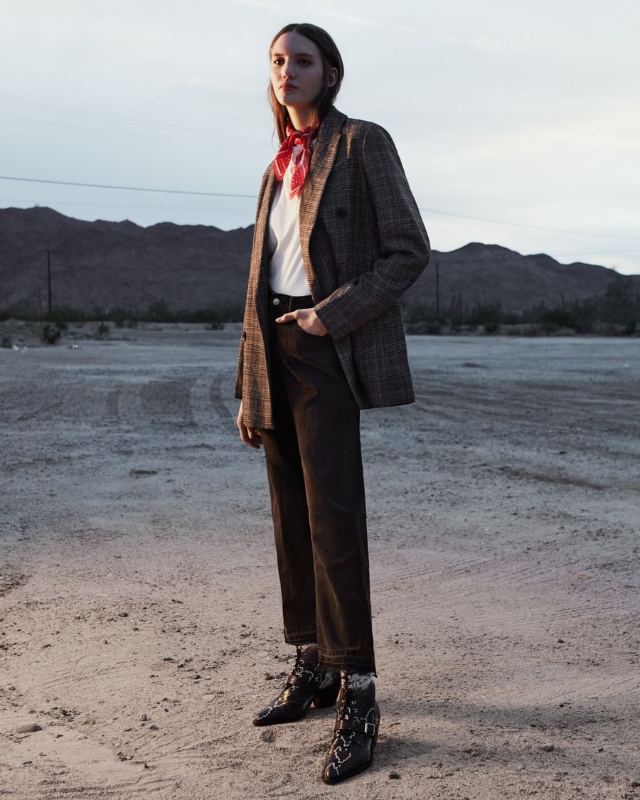 Woman standing in the desert at sunset wearing a white t-shirt with a grey checked blazer, black jeans, snake printed boots and a red scarf around her neck.