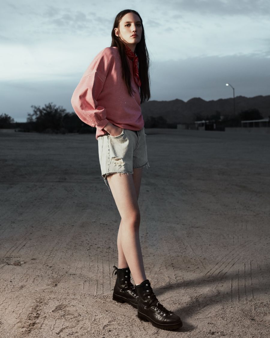 Woman standing in the desert at sunset wearing a light pink sweatshirt with cut off light denim shorts, black leather boots and a red bandana around her neck.